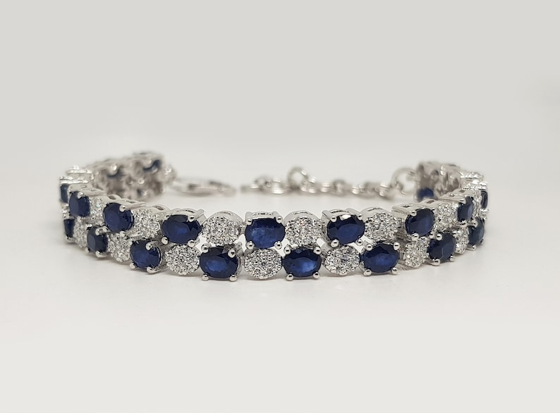 May include: A silver bracelet featuring two rows of oval, dark blue gemstones alternating with smaller, clear stones. The bracelet has a delicate chain closure and is set against a white background.