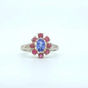 Könnte beinhalten: Ein silberner Ring mit einem zentralen ovalen blauen Edelstein, umgeben von einem Kranz aus kleineren, runden roten Edelsteinen. Der Ring ist vor einem weißen Hintergrund platziert, um die Farben und das Design hervorzuheben.
