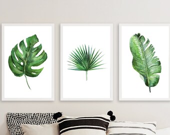 May include: Three framed watercolour prints showcasing green tropical leaves. The prints depict a monstera leaf, a fan palm leaf, and a banana leaf, set against a white backdrop. The artwork is displayed above decorative cushions.
