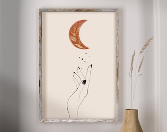 May include: A framed print with a minimalist line drawing of a hand reaching up towards a crescent moon with a gold leaf design. The moon is a rusty orange color against a cream background.