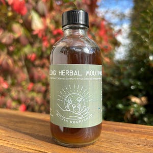 May include: A clear glass bottle with a black cap, filled with a brown liquid. The bottle has a green label with the text "Healing Herbal Mouthwash" and "Wildly Nourished". The background features blurred red and green foliage.
