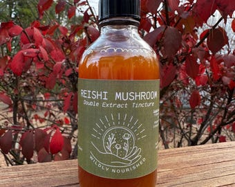 May include: A glass bottle with a black lid, containing an amber liquid. The label reads "REISHI MUSHROOM Double Extract Tincture" with a botanical illustration and the words "WILDLY NOURISHED". The bottle is on a wooden surface, with red leaves in the background.