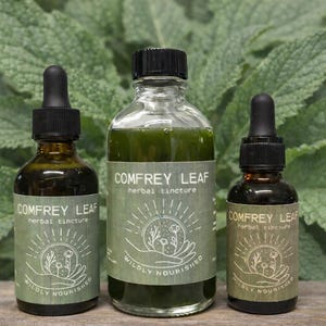 May include: Three glass bottles of Comfrey Leaf herbal tincture with black droppers. The bottles have green labels with the text "COMFREY LEAF herbal tincture" and a graphic of a sun, flowers, and hands. The background is green foliage.