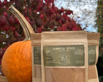 Könnte beinhalten: Ein oranger Kürbis neben drei braunen Papiertüten mit Kürbisgewürzmischung. Die Tüten haben ein grünes Etikett mit dem Text "Pumpkin Spice" und "Autumn Spice Blend". Der Hintergrund zeigt rote und grüne Blätter.