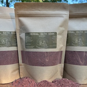 Könnte beinhalten: Drei braune Papiertüten mit Kräuterbädern mit einem transparenten Fenster, das den rotbraunen Inhalt zeigt. Die Beutel haben grüne Etiketten mit dem Text "HERBAL BATH SOAK Sacred Blend" und "WILDLY NOURISHED".