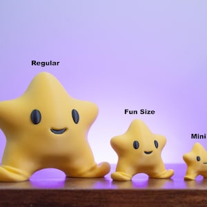 Può includere: Tre figurine a forma di stella gialla di diverse dimensioni, etichettate "Regular", "Fun Size" e "Mini". Ogni stella ha occhi ovali neri e una bocca sorridente. Le figurine sono su una superficie di legno contro uno sfondo sfumato viola.