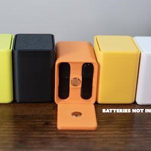 Puede incluir: Una fila de cinco cajas rectangulares coloridas con tapas. Las cajas son amarillas, negras, naranjas, amarillas y blancas. La caja naranja tiene una puerta con bisagras abierta, revelando dos compartimentos oscuros. El texto en la parte inferior dice "BATTERIES NOT INCLUDED."