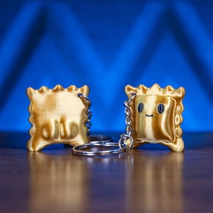 May include: Two gold-colored ravioli-shaped keychains. One keychain has Arabic text, the other has a smiling face. Both keychains have a silver keyring attached. The background is a gradient of blue.