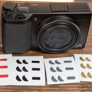 May include: A black digital camera with a lens and a camouflage strap is displayed on a wooden surface. Four sets of stickers with various shapes and colors, including red, black, gray, and gold, are placed below the camera.