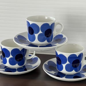 Op de afbeelding: Drie witte keramieken theekopjes met bijpassende schoteltjes, versierd met een opvallend blauw bloemenpatroon. Elke kop heeft een afgerond oortje en een donkerblauw centrum in de bloemblaadjes. De set is opgesteld met één kopje verhoogd.