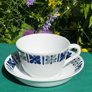 May include: A white ceramic teacup and saucer set featuring a repeating pattern of blue and sage green stylized floral motifs around the exterior of the cup and the rim of the saucer. The cup has a curved handle and a smooth, glossy finish.
