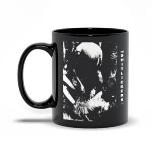May include: Black ceramic coffee mug with a white graphic of a band called "The Shitlickers" with a black and white photo of a person in the background.