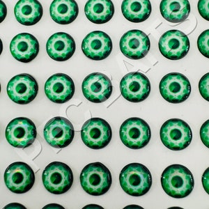May include: A collection of small, round, glass cabochons with a vibrant green and white design. Each cabochon features a central green eye-like pattern surrounded by a white, starburst-like design. The cabochons are arranged in a grid pattern.