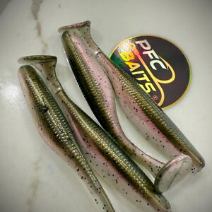 May include: Four soft plastic fishing lures with a fish-like design. The lures are green, pink, and clear with black speckles. A circular sticker with the text "PFC BAITS" is also visible.