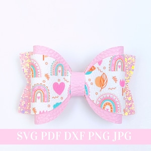 Könnte beinhalten: Eine rosa-weiße Haarschleife mit einem Regenbogen- und Herzmuster. Die Schleife ist aus Kunstleder gefertigt und hat einen Glitzer-Akzent.