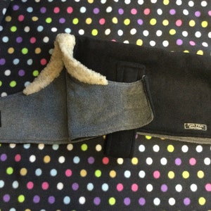 May include: A black fleece dog coat with a gray fleece lining and a white faux fur collar. The coat has a velcro closure and a label that reads "Pooch Papers Foundation".