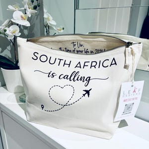 May include: Cream-colored canvas travel bag with the text "SOUTH AFRICA is calling" in black script. A dotted line forms a heart shape with an airplane graphic. The bag has a zipper closure and a tag that says "LEAF DESIGNS MADE JUST FOR YOU."