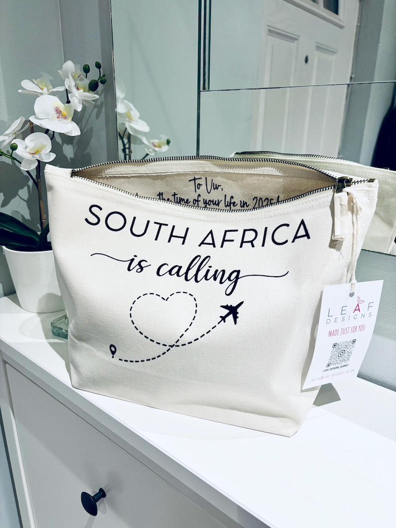 May include: Cream-colored canvas travel bag with the text "SOUTH AFRICA is calling" in black script. A dotted line forms a heart shape with an airplane graphic. The bag has a zipper closure and a tag that says "LEAF DESIGNS MADE JUST FOR YOU."