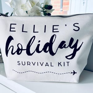 May include: White canvas zippered pouch with black text that reads "Ellie's holiday survival kit" and a small airplane icon.