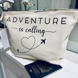 May include: A cream-colored travel pouch with a black zipper and the words "ADVENTURE is calling" printed on the front. A dotted heart and airplane graphic are below the text. A passport rests below the pouch.