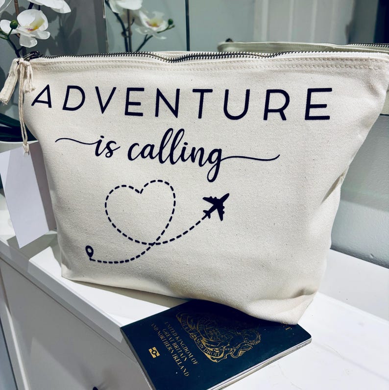 May include: A cream-colored travel pouch with a black zipper and the words "ADVENTURE is calling" printed on the front. A dotted heart and airplane graphic are below the text. A passport rests below the pouch.