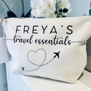 May include: Cream-colored travel essentials bag with the name "FREYA'S" in a bold font, and "travel essentials" in a script font. A dotted heart and airplane graphic are printed on the front. The bag has a zipper closure.