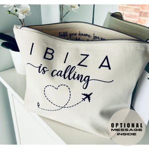 May include: Off-white canvas cosmetic bag with the words "IBIZA is calling" in black script. A dotted line with a heart and airplane graphic is below the text. The bag has a black zipper and the words "Optional Message Inside" are printed on the bottom right.