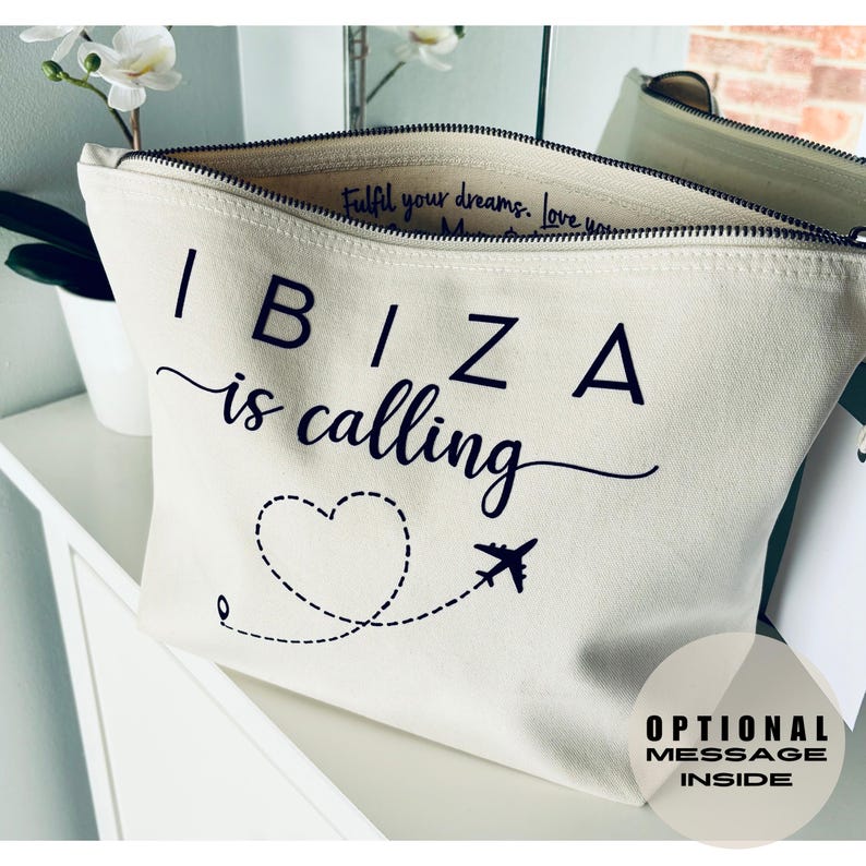 May include: Off-white canvas cosmetic bag with the words "IBIZA is calling" in black script. A dotted line with a heart and airplane graphic is below the text. The bag has a black zipper and the words "Optional Message Inside" are printed on the bottom right.