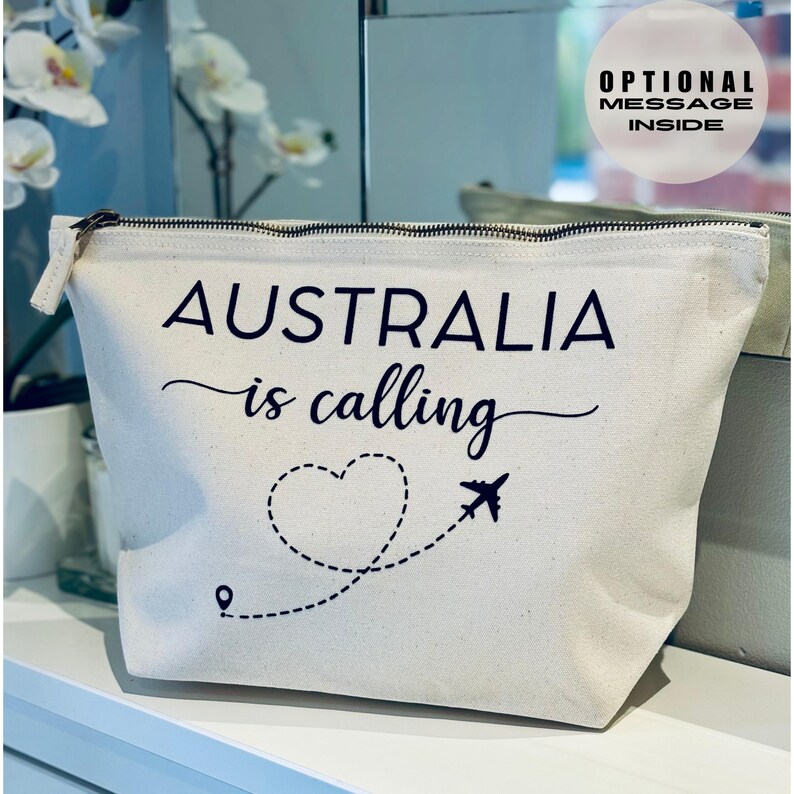 May include: Cream-colored travel pouch with a black zipper and the phrase "AUSTRALIA is calling" in black script. A heart-shaped dotted line with a plane graphic is below the text. The pouch has a rectangular shape.