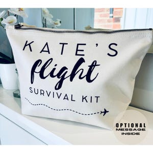 May include: A white canvas zippered pouch with black text that reads "KATE'S flight SURVIVAL KIT" and a small airplane icon. The pouch has a dashed line and an arrow pointing to the right.