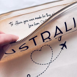 May include: Cream-colored travel pouch with a gold zipper, featuring the word "AUSTRALIA" in large black letters. A dotted line with a heart and airplane graphic is visible. The text "To Alice, you were made for this... Love from S..." is also printed.