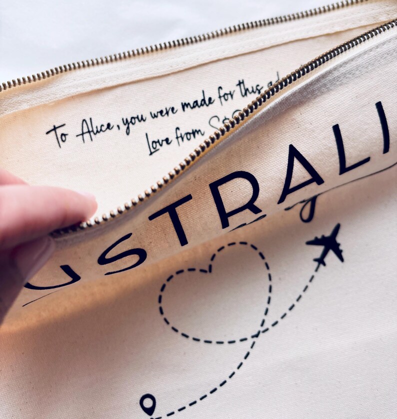 May include: Cream-colored travel pouch with a gold zipper, featuring the word "AUSTRALIA" in large black letters. A dotted line with a heart and airplane graphic is visible. The text "To Alice, you were made for this... Love from S..." is also printed.