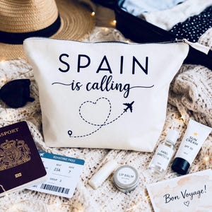 May include: Cream-colored travel pouch with the text "SPAIN is calling" and a heart-shaped dotted line with an airplane graphic. Accessories include a passport, boarding pass, sunglasses, lip balm, hand sanitizer, and sunscreen.