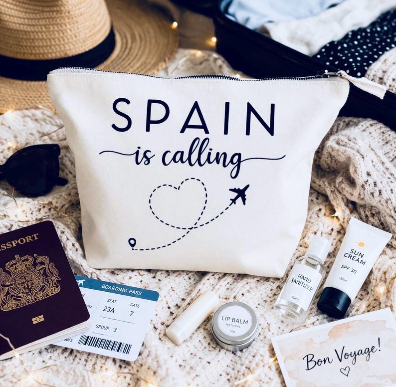 May include: Cream-colored travel pouch with the text "SPAIN is calling" and a heart-shaped dotted line with an airplane graphic. Accessories include a passport, boarding pass, sunglasses, lip balm, hand sanitizer, and sunscreen.