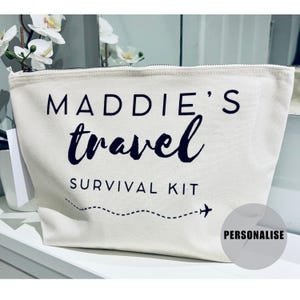 May include: A white canvas zippered pouch with black text that reads "MADDIE'S travel SURVIVAL KIT" with a dotted line and airplane graphic. The pouch has a grey "PERSONALISE" label.