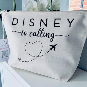May include: A cream-colored zippered pouch with the text "DISNEY is calling" in black. A dotted line forms a heart shape and leads to a small airplane graphic. The pouch has a tag attached.