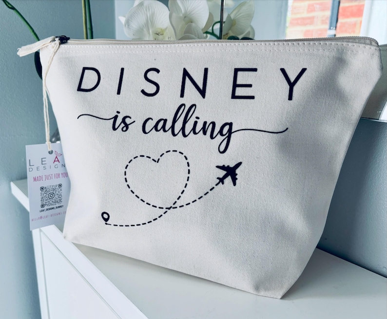 May include: A cream-colored zippered pouch with the text "DISNEY is calling" in black. A dotted line forms a heart shape and leads to a small airplane graphic. The pouch has a tag attached.