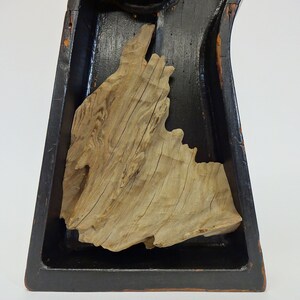 May include: A natural wood sculpture with a light brown, textured surface, set within a black, rectangular frame. The wood grain is visible, and the sculpture has an organic, irregular shape, suggesting a piece of driftwood or a similar natural element.