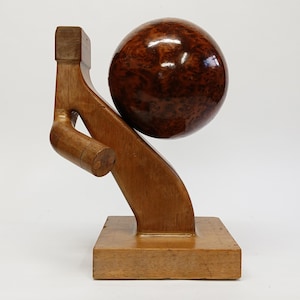May include: A wooden sculpture featuring a sphere with a burl wood pattern, held by a stylized wooden arm and base. The sculpture is made of light brown wood and has a smooth, polished finish. The sphere is a rich, dark brown color.