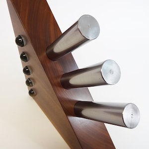 Puede incluir: Una estantería triangular de madera con tres varillas de metal plateado fijadas en la parte delantera. La estantería tiene cinco detalles redondos de vidrio negro y transparente en el lateral.