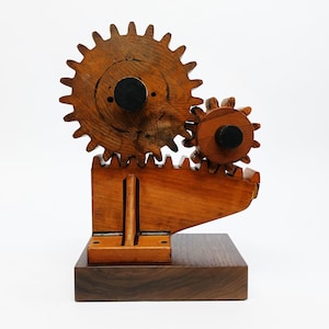 May include: Wooden gear sculpture with two interlocking gears of different sizes. The gears are made of light brown wood and mounted on a dark brown wooden base. The sculpture has a rustic, handcrafted appearance.