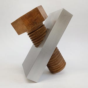 May include: A modern sculpture featuring a large wooden bolt intersecting a brushed metal rectangular block. The bolt has a square head and threaded shaft. The sculpture is set against a white background.