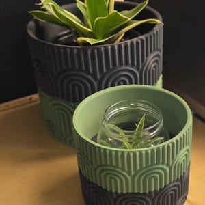 Peut inclure: Deux pots de fleurs décoratifs, un grand et un petit, avec un motif géométrique. Le grand pot est noir et vert, contenant une plante verte et jaune. Le petit pot est vert et noir, avec un bocal en verre à l'intérieur.
