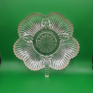 May include: Clear glass serving dish with a unique, scalloped, leaf-like shape. The dish features a central, circular design with radiating lines and starburst patterns. The edges have a textured, ridged design, and a small footed base.