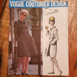 May include: Vogue Couturier Design pattern number 1427 by Simonetta for a coat. The pattern shows two models wearing different versions of the coat. One model is wearing a grey coat with a white hat and the other is wearing a white coat with a yellow hat.