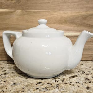 May include: A white ceramic teapot with a simple design. The teapot has a rounded body, a spout, a handle, and a lid.