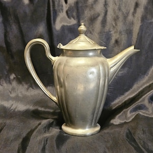 May include: A vintage silver-colored teapot with a curved handle, a spout, and a lid with a decorative finial. The teapot has a ribbed design on its body and sits on a small, round base. The background is a dark, textured fabric.