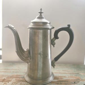 May include: A silver coffee pot with a black handle. The pot has a rounded body and a lid with a finial. The pot is sitting on a wooden surface.