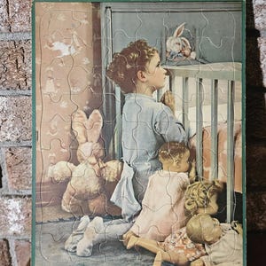 May include: A vintage jigsaw puzzle depicting a young girl kneeling in prayer beside a crib with a baby doll and a stuffed bunny. The girl is wearing a blue dress and has her hands clasped together. The crib is white with a white blanket and a stuffed bunny on top. The background is a light brown wallpaper with a floral pattern.