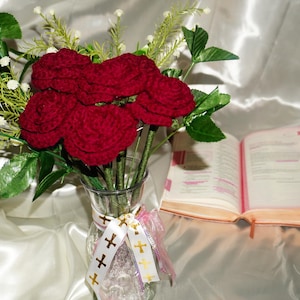 May include: A bouquet of deep red crocheted roses with green stems and leaves arranged in a clear glass vase. The vase is decorated with a white ribbon featuring gold cross symbols. An open book with pink page markers sits beside the vase.